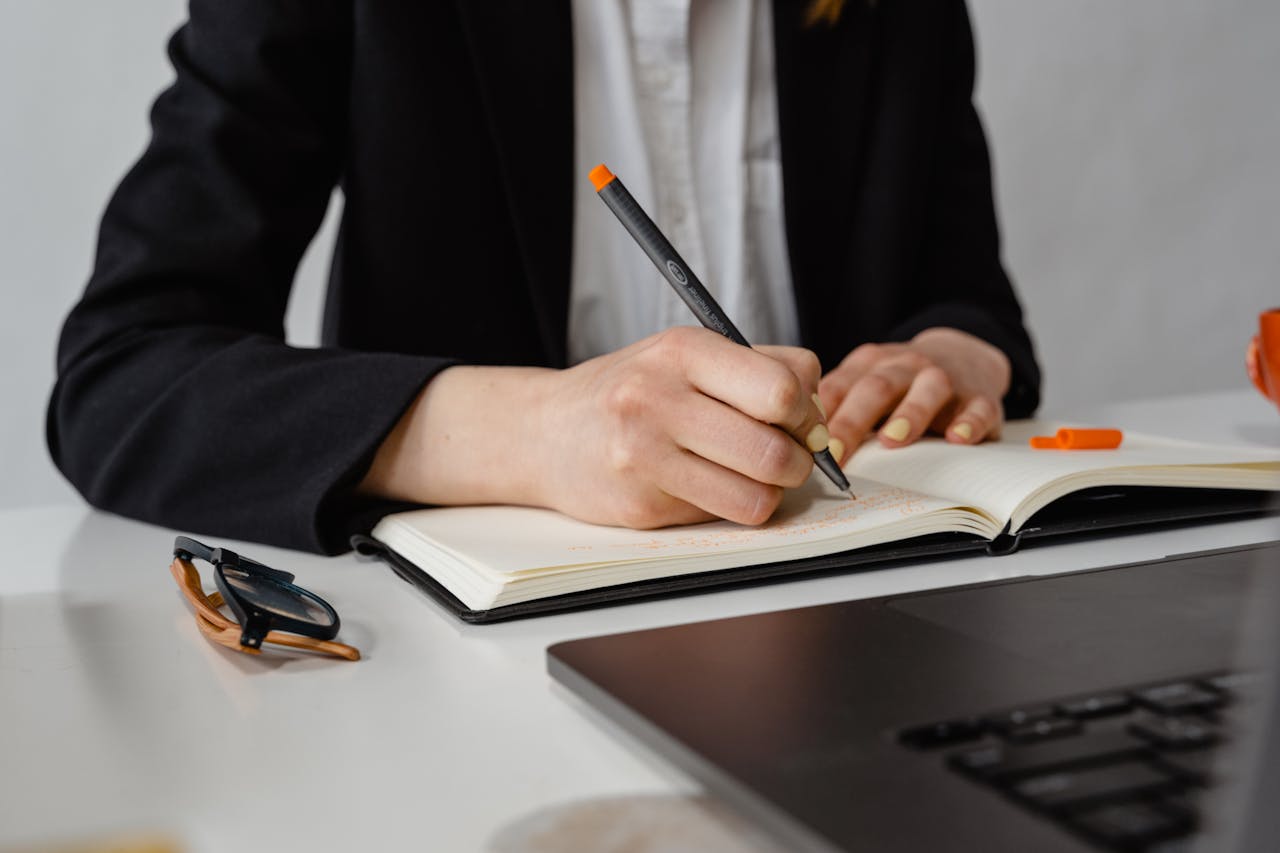Person writing notes in a planner on a desk, emphasizing a professional and organized workspace.